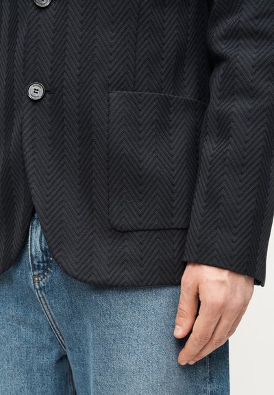 Blazer noir ajusté à motif chevrons, avec deux boutons et une poche avant, associé à un jean en denim bleu clair.