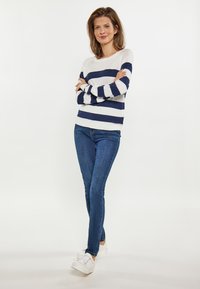 Gestreifter Strickpullover in Weiß und Marineblau, kombiniert mit blauen Jeans und weißen Sneakers. Das Model steht mit verschränkten Armen vor einem neutralen Hintergrund.