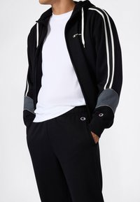 Uomo che indossa una felpa nera con zip, strisce bianche e toppe grigie sui gomiti, una maglietta bianca e pantaloni della tuta neri con logo sulla tasca.