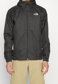 Veste noire imperméable avec une fermeture éclair frontale, des poignets élastiques et le logo The North Face sur la poitrine. Texture lisse et capuche.