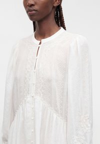 Femme aux cheveux tressés portant un chemisier blanc boutonné présentant des motifs texturés complexes et un col rond.