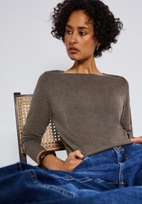 Khakifarbener Pullover mit langen Ärmeln und Bootsausschnitt, weicher Textur, kombiniert mit blauen Jeans, sitzt in einem geflochtenen Holzstuhl.