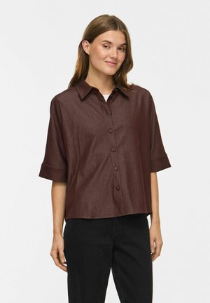 Femme aux cheveux châtain clair portant une chemise marron foncé ample à manches courtes avec des boutons et un pantalon noir sur un fond uni.