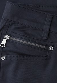 Gros plan sur un pantalon noir avec une poche zippée diagonale, une tirette de fermeture éclair en métal, une boucle de ceinture et des détails de couture visibles.
