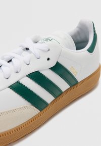 White leather sneaker with green synthetic stripes, gum sole, and textured accents. Features lace-up closure and branded tongue label.