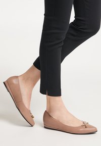 Beige bőr balettcipők apró aranyszínű masnis részletekkel viselve fekete slim-fit nadrággal egy világos háttér előtt.