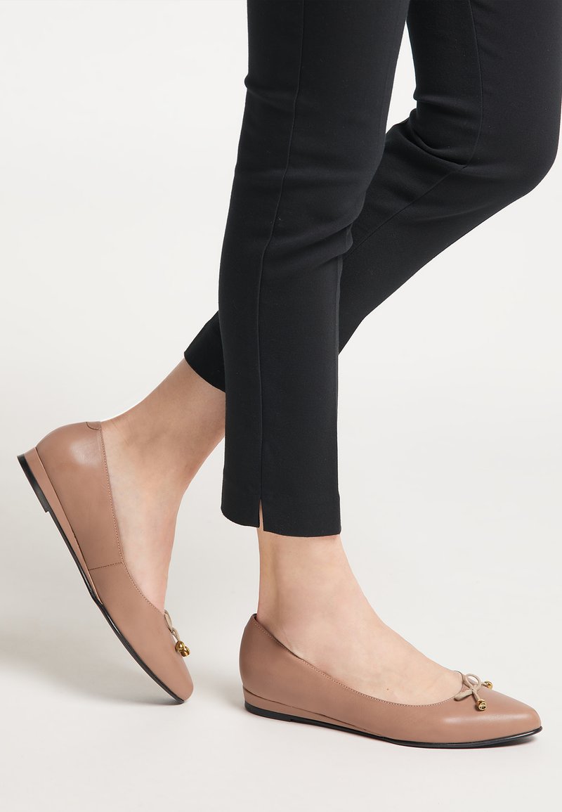 Beige bőr balettcipők apró aranyszínű masnis részletekkel viselve fekete slim-fit nadrággal egy világos háttér előtt.