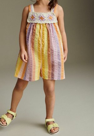 Une combinaison rayée dans des couleurs pastel (jaune, rose, lavande) avec un tissu à volants et un empiècement en dentelle comportant des appliqués de fleurs. Portée avec des sandales jaunes.