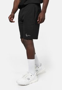 Pantalones cortos de algodón negros con un ajuste relajado, que cuentan con bolsillos laterales y una marca bordada en blanco en el dobladillo. Combinados con zapatillas y calcetines blancos.