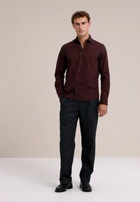 Bordo spalvos ilgomis rankovėmis marškiniai su sagomis ant apykaklės, derinami su tamsiomis kelnėmis ir juodais batais, fone neutralios spalvos.