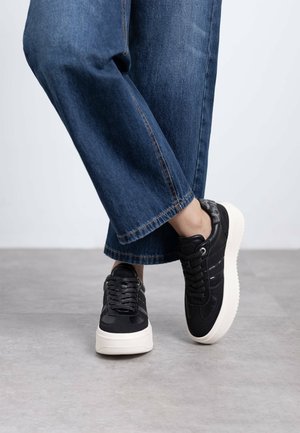 Zapatillas negras de cordones con parte superior texturizada, que presentan detalles en gris y una suela blanca gruesa, combinadas con pantalones de mezclilla anchos.
