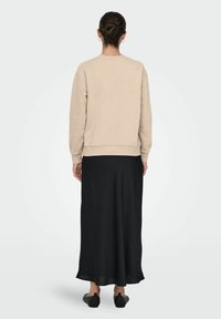 Femme avec les cheveux en chignon portant un pull à manches longues beige, une jupe noire jusqu'aux chevilles et des chaussures noires sans lacets, debout de dos sur un fond uni.