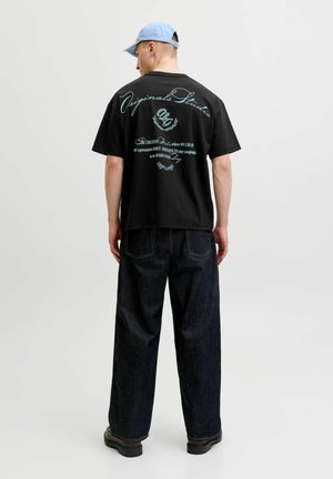 Homme portant un t-shirt graphique noir, un jean large foncé, des chaussures marron et une casquette bleu clair, debout dos à un fond uni.