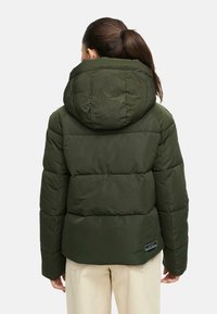 Femme avec une queue de cheval brune portant une veste rembourrée olive avec capuche et un pantalon beige, de dos contre un fond uni.