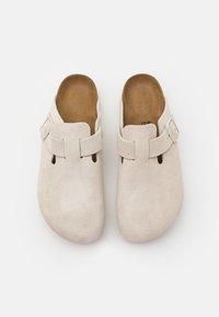 Beige suède clogs met een afgeronde neus, open achterkant, verstelbare band en twee ventilatiesleuven aan elke kant. Glad binnenvoetje.