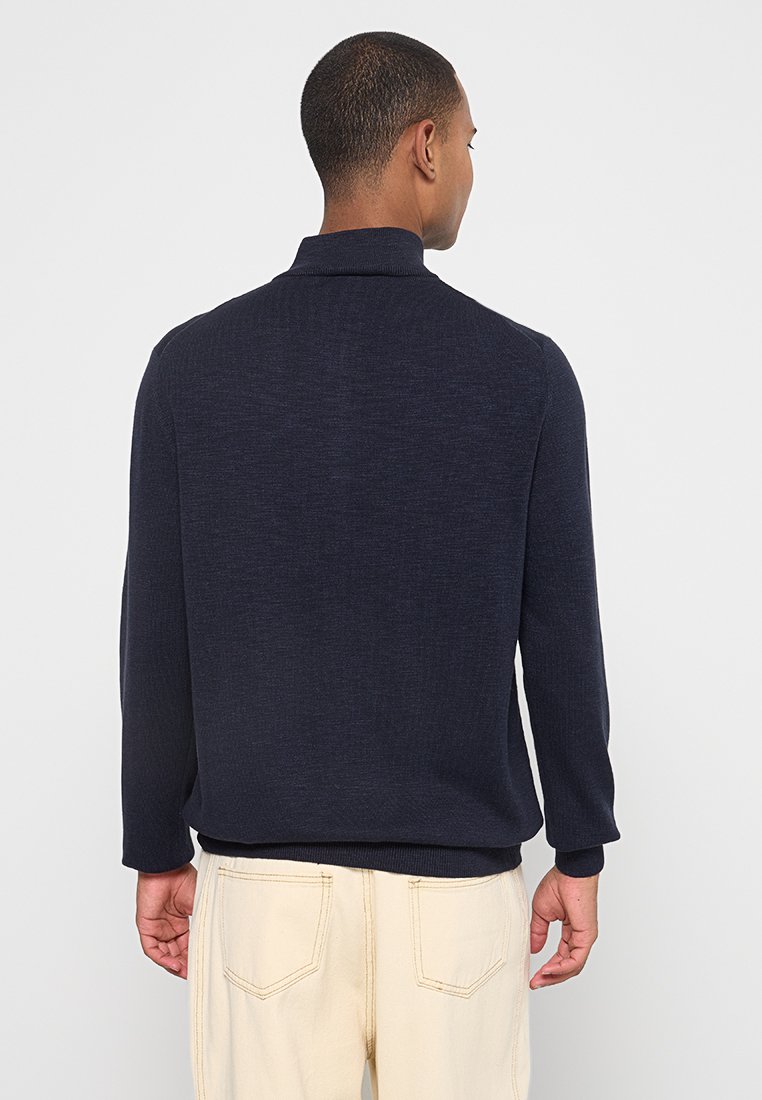 Mężczyzna w ciemnogranatowym swetrze z długim rękawem i jasnobeżowych spodniach, stojący tyłem z rękami luźno wystawionymi wzdłuż ciała.