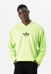 Neon zelena majica s dugim rukavima s teksturiranim mrežastim dizajnom, koja ima crni Adidas logo u središtu i oblik V-izreza.