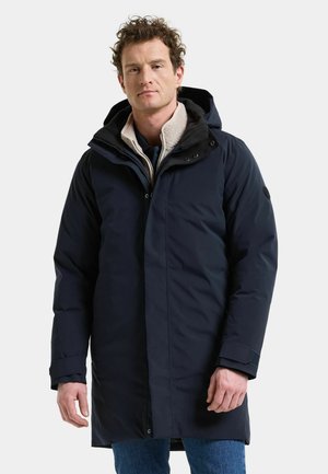 Man som bär en marinblå lång vinterrock med huva och beige innerfoder i fleece, står mot en slät ljus bakgrund.
