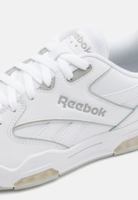 Fehér bőrsnekkerek hálós részletekkel, gumitalppal és ezüst Reebok logóval. Jellemzői között szerepelnek a fűzők és egy látható légpárnás egység.