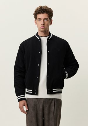 Jeune homme aux cheveux bouclés portant une veste varsity noire sur un pull blanc et un pantalon marron à carreaux, debout devant un fond clair uni.