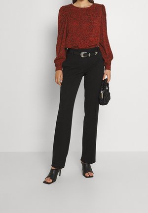 Vrouw draagt een rode blouse met patroon, zwarte hoge taillebroek met een zilveren gesp, zwarte muiltjes met hakken, en houdt een kleine zwarte handtas vast.