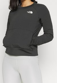 Vrouw in een zwart North Face sweatshirt met lange mouwen, met haar handen in de voorzak en lichte grijze legging.