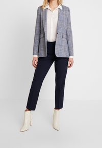 Rutig kavaj i ljusblått och grått, med två framfickor, bärs över en vit skjorta. Kombineras med marinblå slim-fit byxor och krämfärgade ankelstövlar.