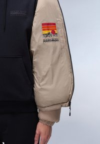Giacca puffer beige con zip nera, con una felpa con cappuccio blu navy sotto. La manica sinistra presenta un logo colorato di un tramonto montano.