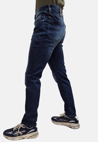 Jeans in denim blu scuro con vestibilità slim, cinque tasche e vita media. Presentano cuciture arancioni e una toppa in pelle sulla cintura posteriore.