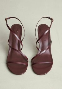 Een paar bruine lederen sandalen met dunne kruislings straps en enkelgespen, geplaatst op een lichtbeige oppervlak.
