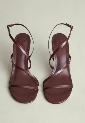 Et par brune lædersandaler med tynde krydssnore og ankelspænder, placeret på en lys beige overflade.