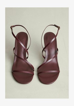 Biezi brūni ādas čības ar plānām krustotu siksnām un potīšu sprādzēm, novietotas uz gaiši bēša virsmas.