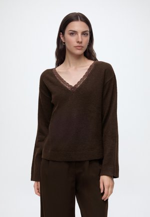 Ung kvinde med langt mørkt hår iført en mørkebrun V-hals sweater med blondekant og matchende mørkebrune bukser mod en ensfarvet baggrund.