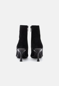 Botas de tornozelo em camurça preta com um alto salto stiletto, apresentando um design elegante e um detalhe texturizado ao longo do lado. Fecho de correr na parte de trás.