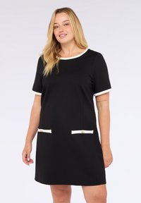 Vestido negro de manga corta con ribete blanco, dos bolsillos frontales con botones dorados y un escote redondo, hecho de una tela suave.