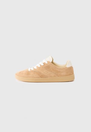 Sneaker in suede color sabbia con lacci e dettagli bianchi, caratterizzato da una superficie testurizzata, punta arrotondata e una suola in gomma con battistrada a pattern.