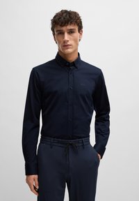 Marineblaue Hemdbluse mit langen Ärmeln, figurbetontem Design, dezentem Branding auf der Brust, kombiniert mit strukturierten dunklen Hosen.