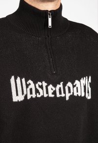 Melnā adīta džemperis ar augstu apkakli, priekšpusē ar balto drukāto logo "Wastedparts" un pusi-izjūgu.