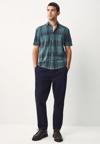 Kortærmet ternet skjorte i teal med mørkegrønne og lilla accenter, kombineret med løstsiddende navy bukser og brune sko.