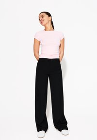 T-shirt ajustée rose clair, pantalon noir à jambes larges et baskets blanches. Modèle se tenant avec les mains derrière le dos, légèrement tourné vers la droite.