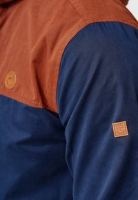 Épaule de veste avec un panneau supérieur couleur rouille, un panneau inférieur et une manche bleu marine, avec deux petites étiquettes de marque en cuir tanné.