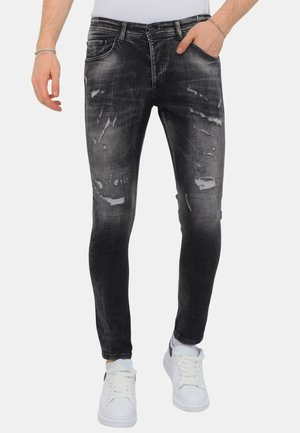 Man draagt smalle zwarte jeans met slijtage-effect, witte sneakers en een wit shirt, één hand in de zak, staand tegen een witte achtergrond.