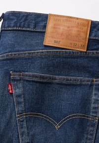 Vaqueros de mezclilla azul con etiqueta de cuero marrón que dice "LEVI STRAUSS & CO." y "501" en dorado. Costura amarilla y pestaña roja en el detalle del bolsillo.
