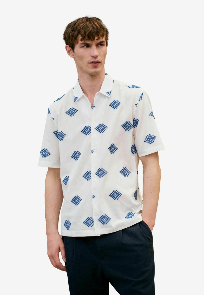 Camisa de manga corta blanca con patrones geométricos azules, confeccionada en tejido ligero; presenta un cuello abotonado y un corte relajado.