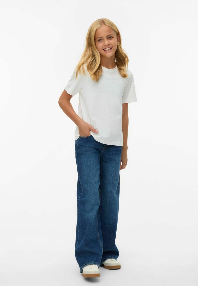 T-shirt blanc à manches courtes rentré dans un jean large bleu, de coupe droite. Le mannequin porte des baskets blanches avec une semelle beige.