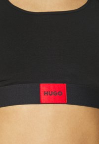 Čierna športová podprsenka so priliehavým strihom a zaobleným výstrihom. Na spodnej časti sa nachádza červený elastický pás s označením "HUGO".