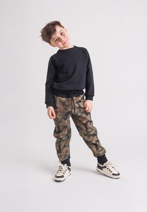 Chłopiec ubrany w czarną bluzę i spodnie moro, pochylający głowę z lekkim uśmiechem, stojący na jednolitym białym tle.