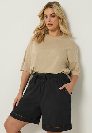 Kvinde med blond hår iført en beige løs t-shirt og sorte teksturerede shorts med elastisk talje og sidelommer, poserer mod en lys baggrund.