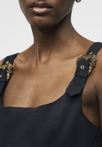 Κοντινό πλάνο ενός ατόμου που φοράει σκούρο, χωρίς μανίκια ένδυμα με περίτεχνες χρυσές πόρπες στα λουριά.