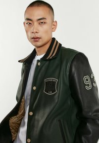 Veste universitaire en cuir vert avec manches noires, col côtelé à rayures et patch brodé sur la poitrine. Dispose de boutons-pression et de poches latérales.